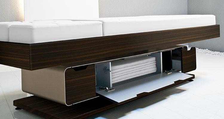 Luxurious Gharieni spa treatment table with sleek wooden design and towel storage.
