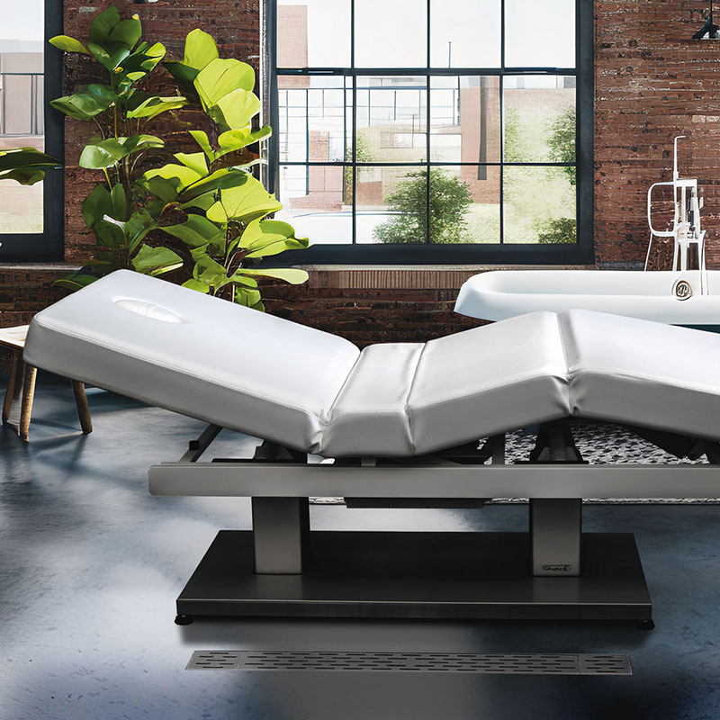 Gharieni premium spa treatment table in modern loft setting with lush greenery.